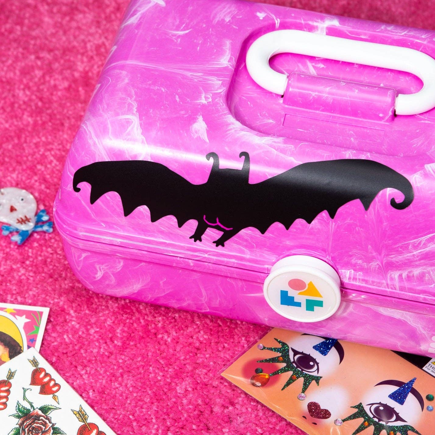 A pink plastic first-aid style first-aid kit with a black bat silhouette decal and a white handle, shown on a textured hot pink surface with small decorative cards nearby.
