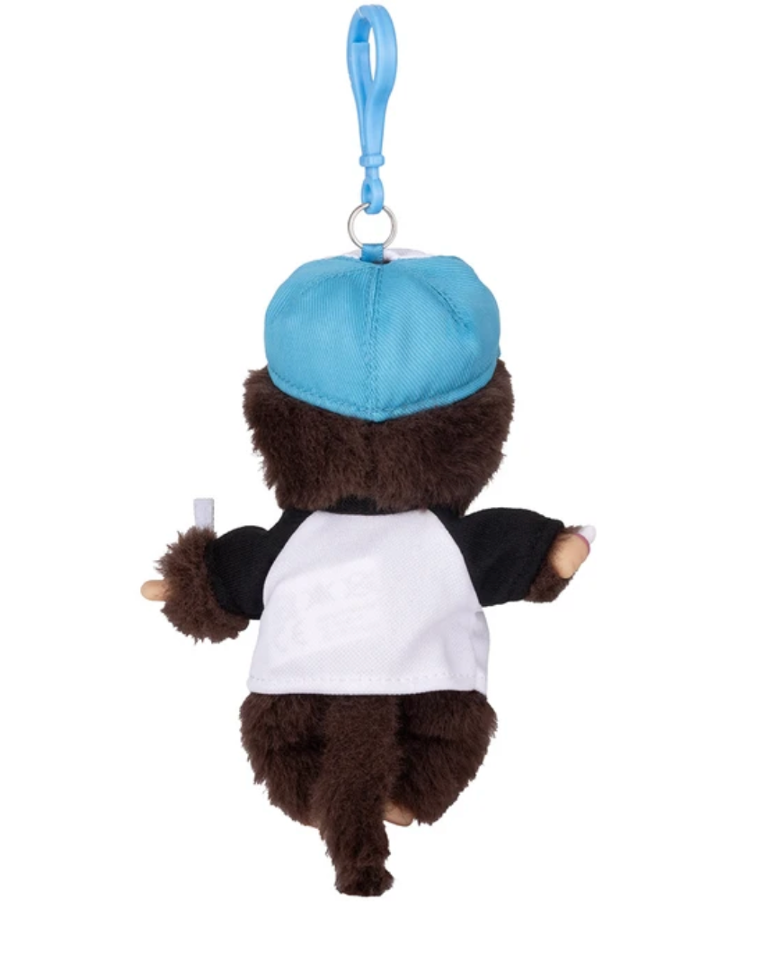 A plush toy keychain or bag charm shaped like a small brown bear wearing a light blue cap, a white and black rag-style shirt, and holding a small white item, shown against a plain white background.