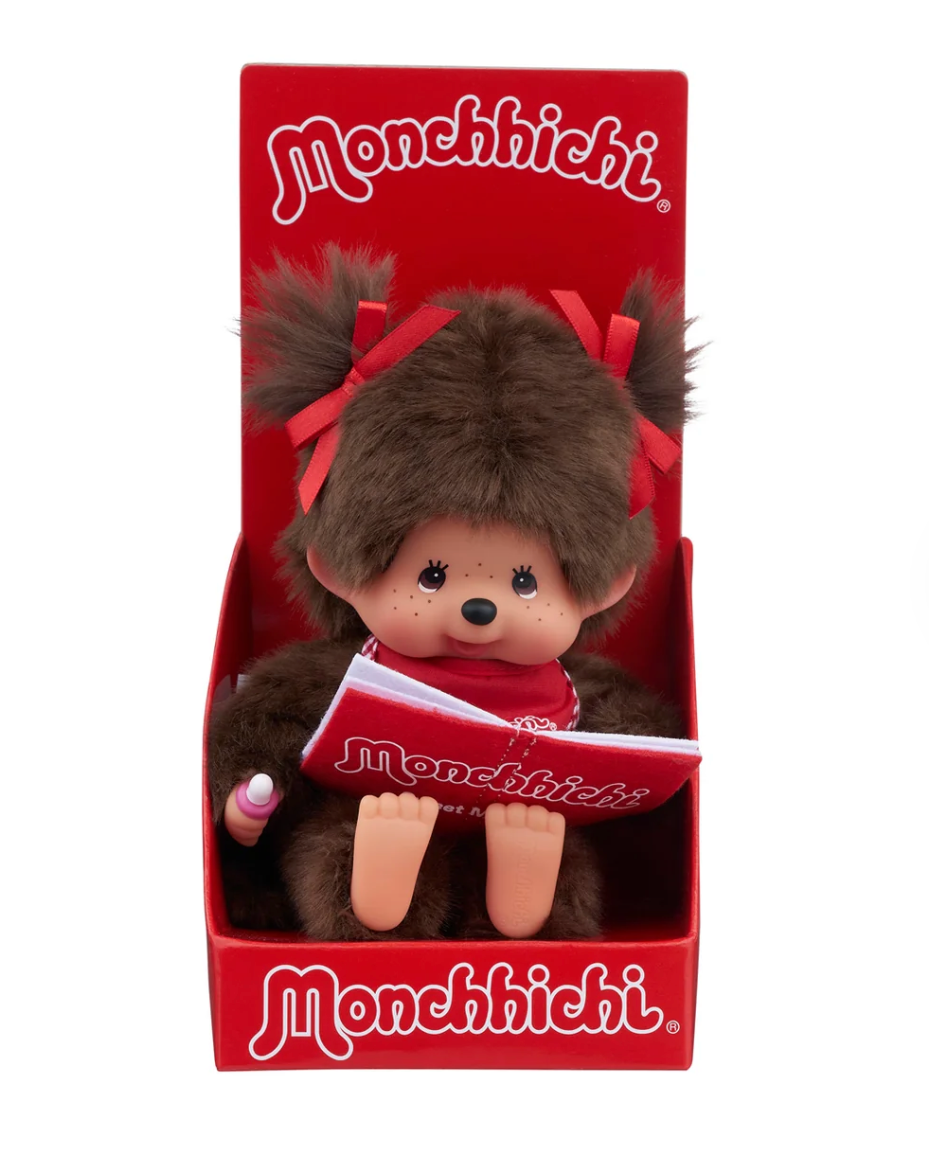 A Monchhichi plush toy doll sitting inside its red retail display box, holding a small Monchhichi book. The doll has brown faux fur, red bows in its hair, and a red outfit.