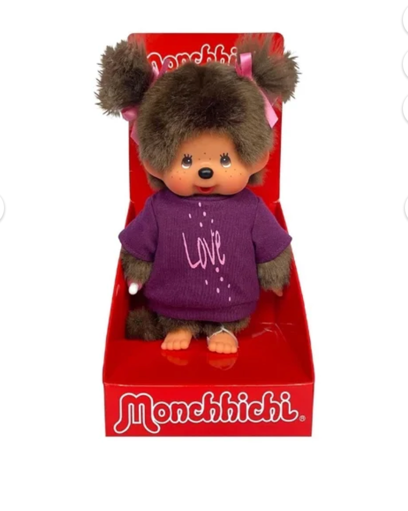 A Monchhichi plush toy doll with brown fur, pink hair bows, and a purple sweater, displayed inside its red retail packaging box against a white background.