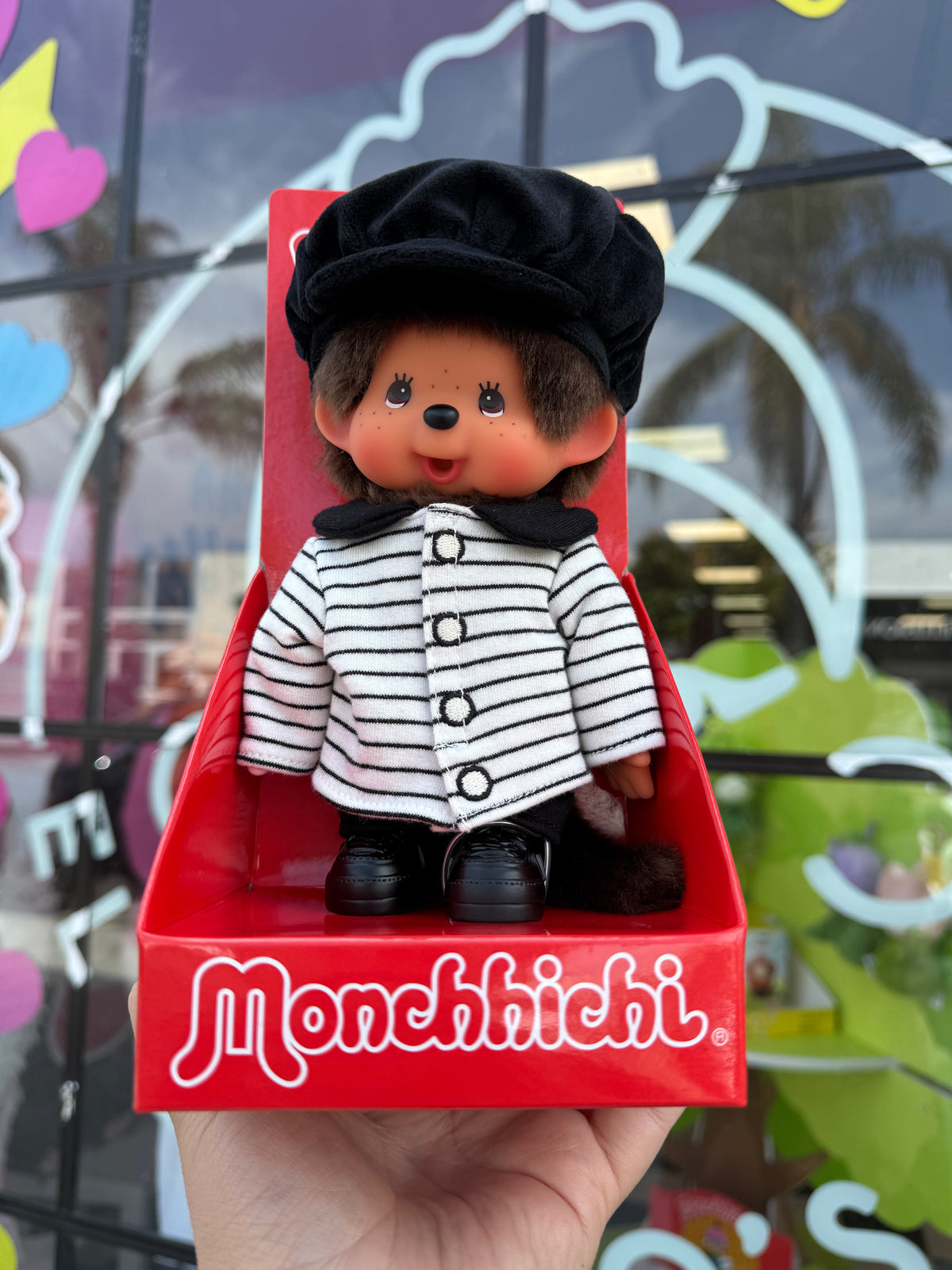 A Monchhichi doll dressed in a black beret, black-and-white striped jacket, and black shoes, displayed in a red Monchhichi box held in a hand. The background shows a colorful, blurred storefront window.