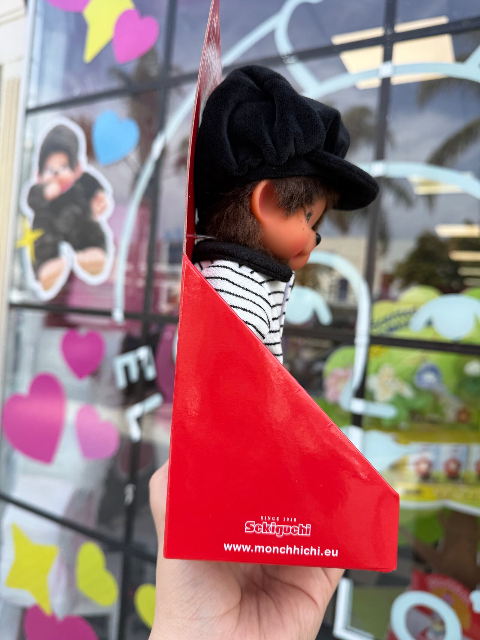 A Monchhichi doll dressed in a black beret, black-and-white striped jacket, and black shoes, displayed in a red Monchhichi box held in a hand. The background shows a colorful, blurred storefront window.