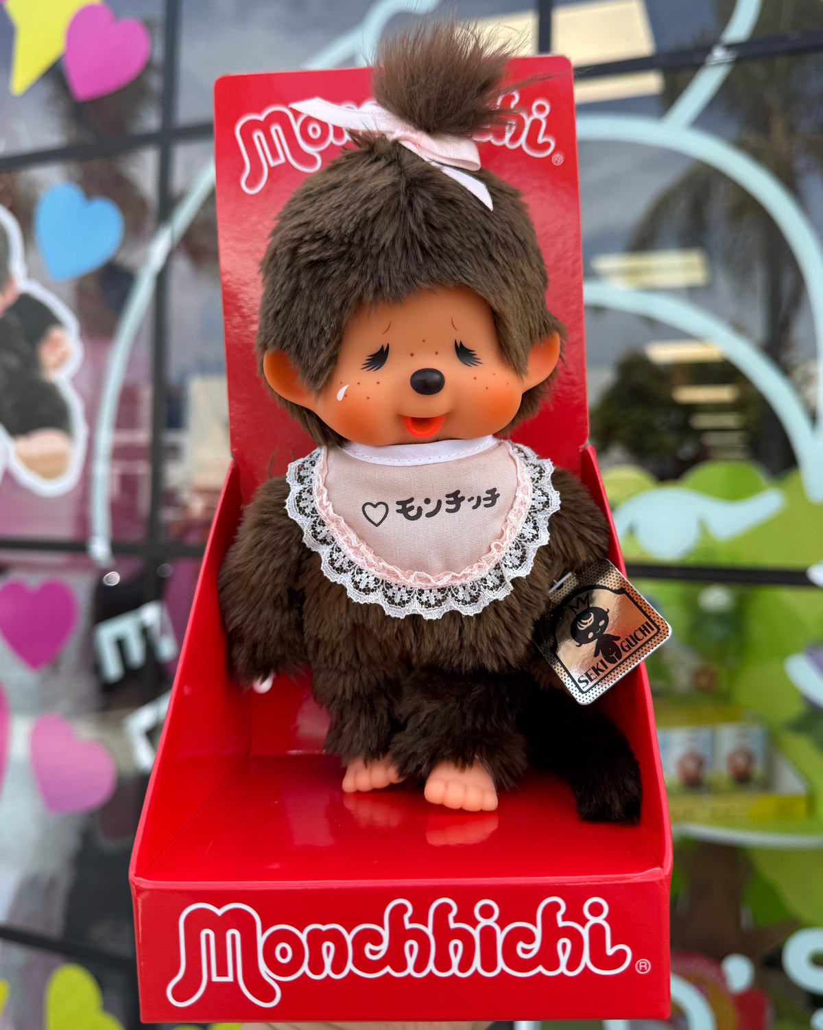 A Monchhichi plush toy doll with brown fur, a pink bow, and a white bib, displayed inside its red retail box held by a hand. The background shows a colorful, slightly blurred storefront window.