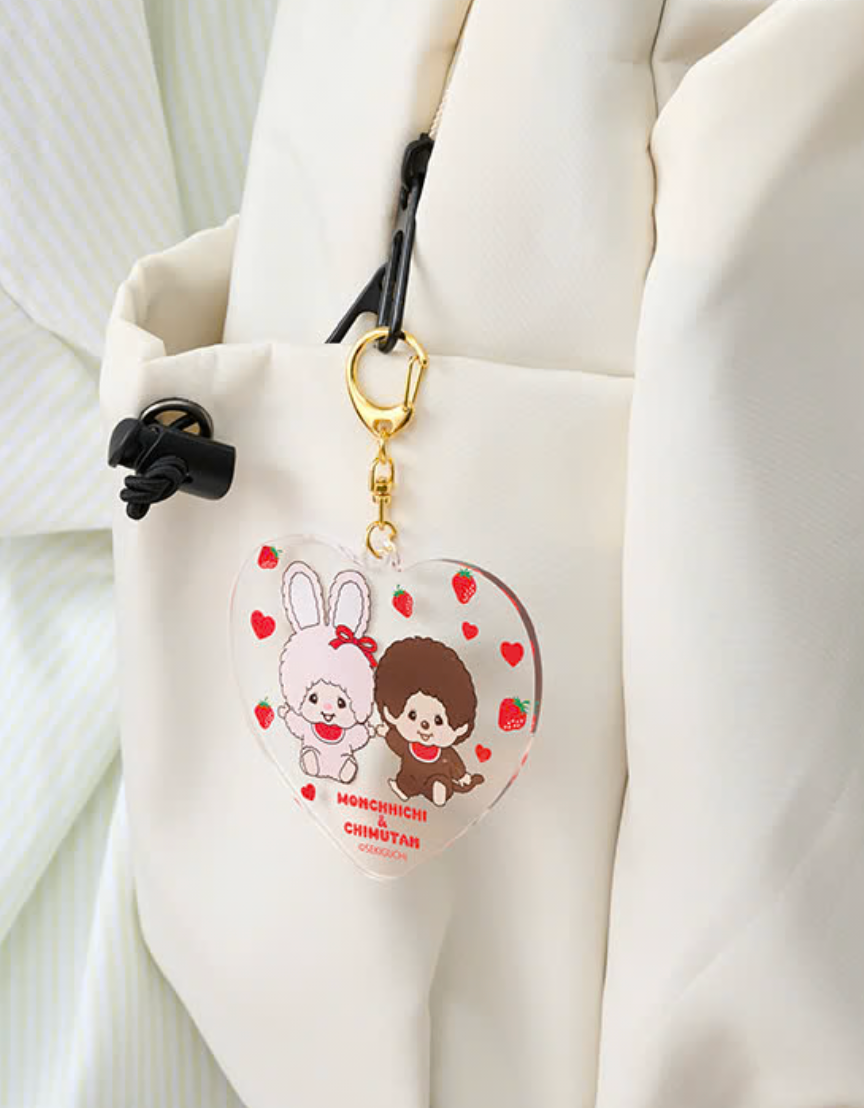 A heart-shaped acrylic keychain charm featuring a cute cartoon bunny and a small character with strawberries, attached to a gold clasp and black strap, shown hanging on a white bag.