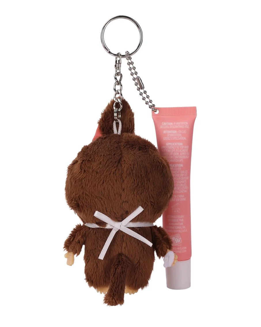A brown plush keychain shaped like a small character with a white bow, attached to a silver chain, shown next to a pink cosmetic tube on a plain white background.
