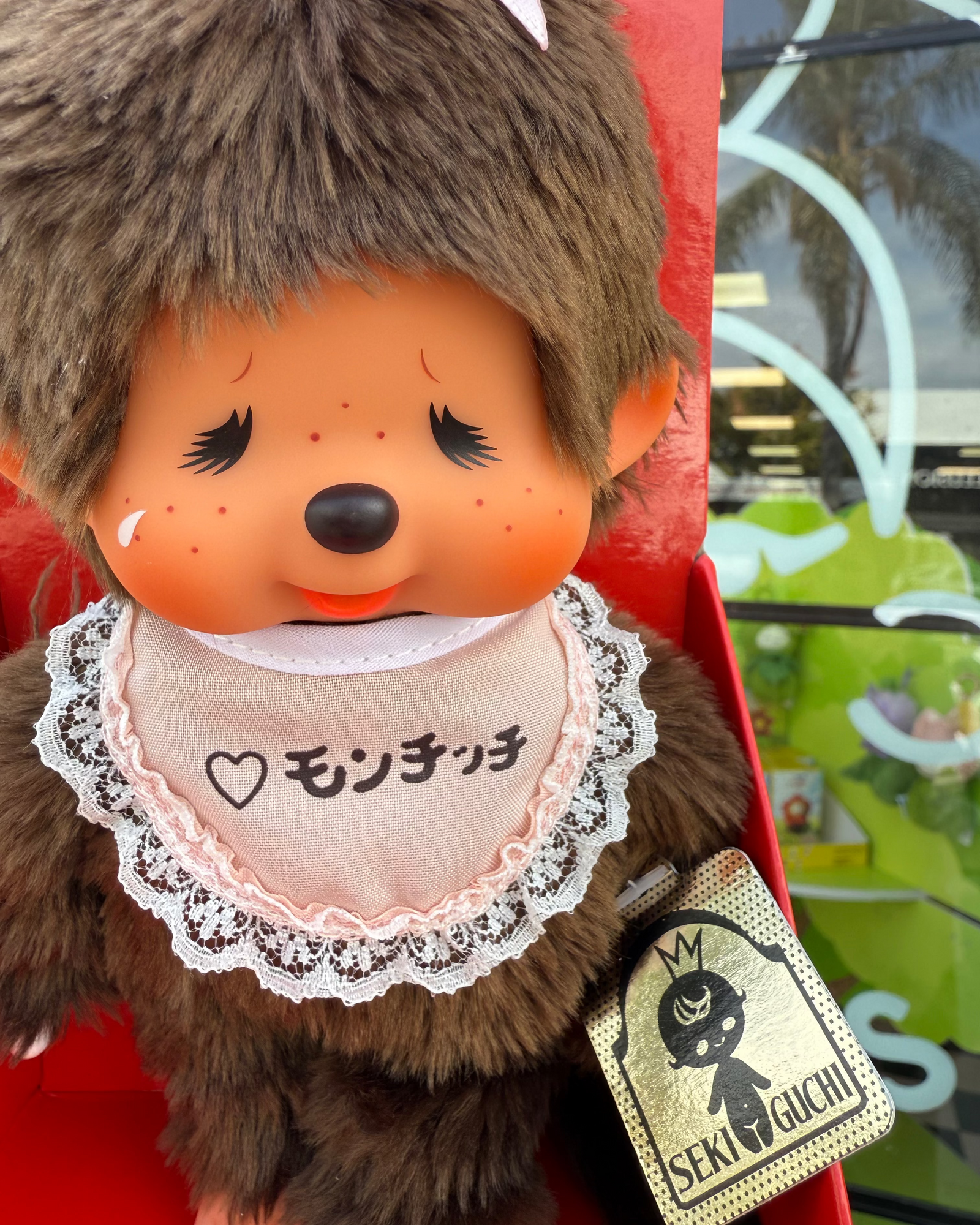 A close-up of a brown plush monkey doll with a peach face, black eyelashes, freckles, and a cream bib printed with Japanese text. The doll is seated in a red display box, with a small gold tag attached.