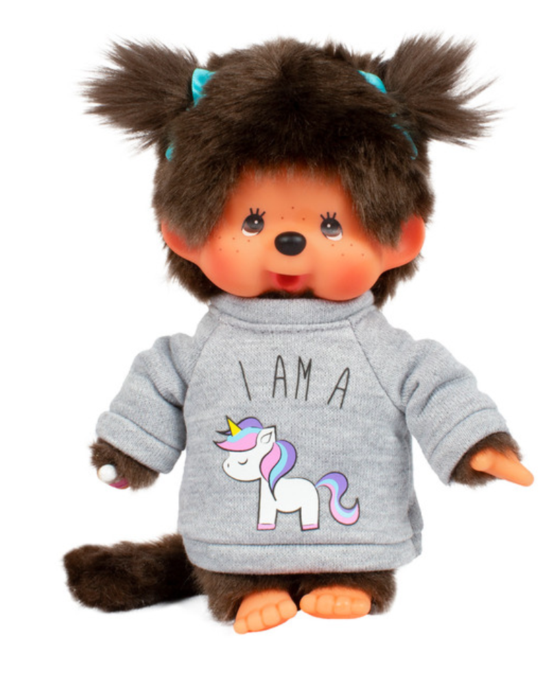 A Monchhichi doll with a unicorn themed grey sweater, featuring its name 'I AM A' on the front. The doll has brown hair, blue hair ties, and a brown base.